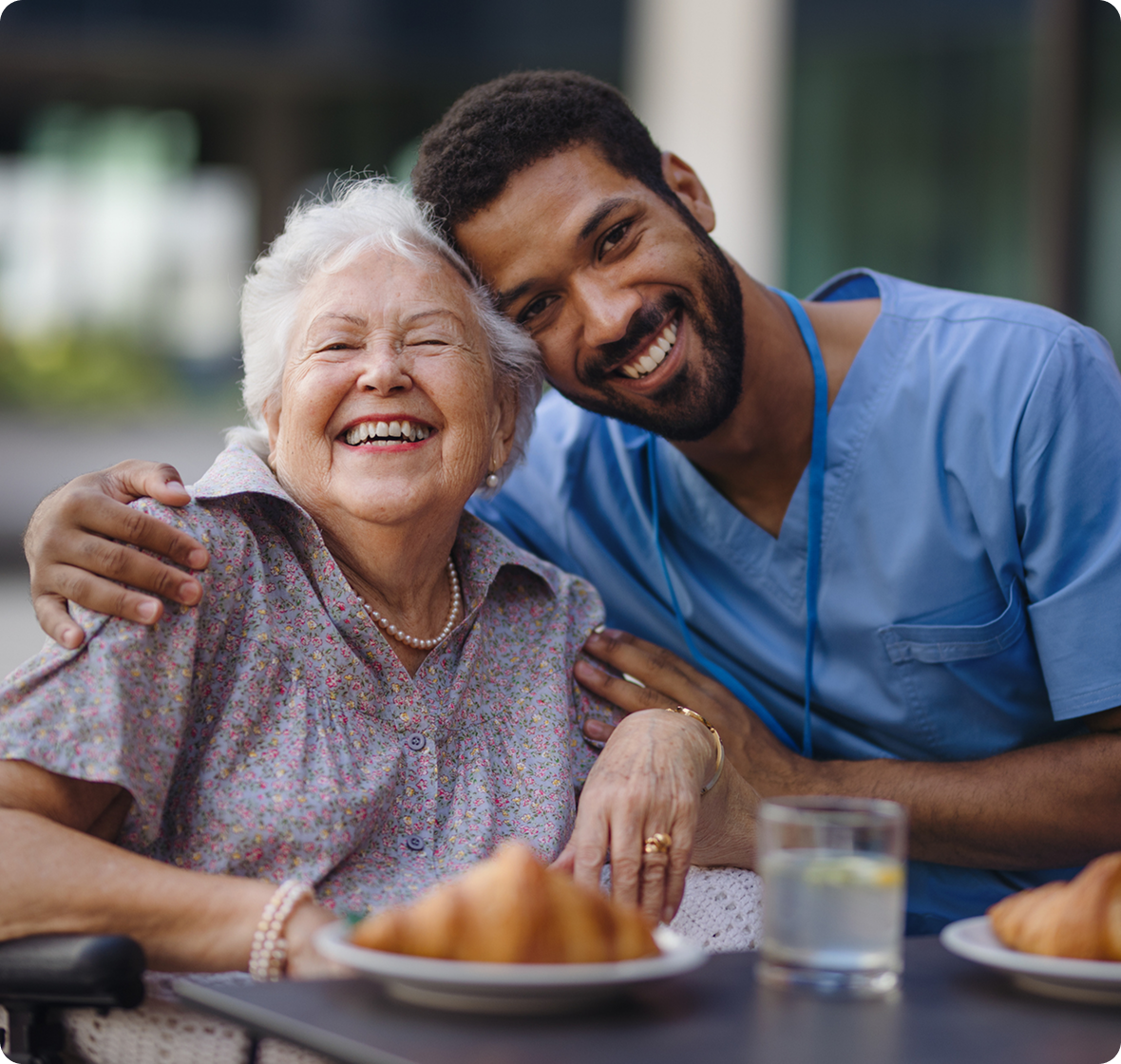Smiling elderly woman embraces a male caregiver in blue scrubs at a dining table with pastries and drinks, conveying joy and companionship.