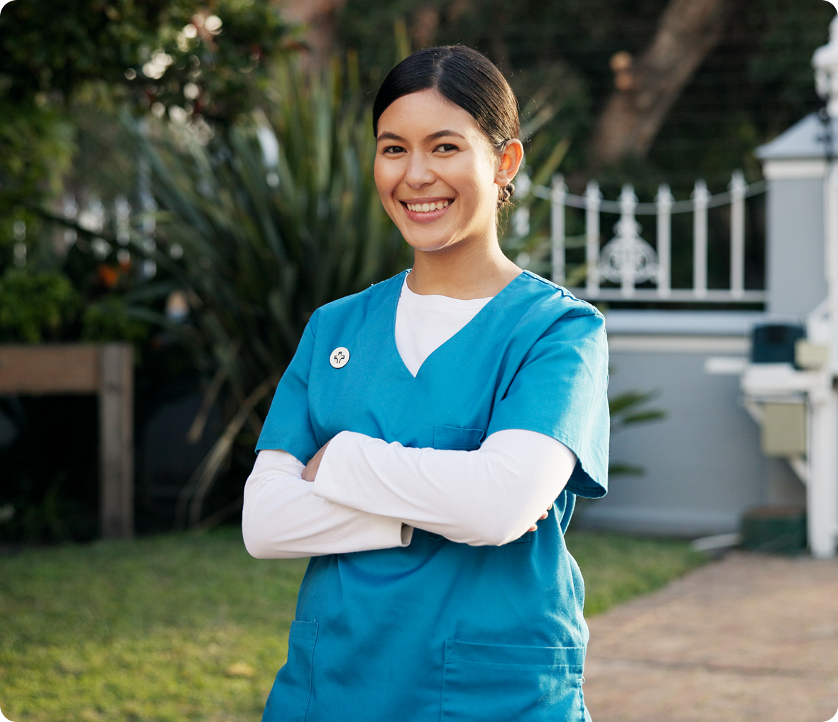 Smiling healthcare worker in blue scrubs stands outdoors with arms crossed, conveying warmth and confidence in a garden setting.
