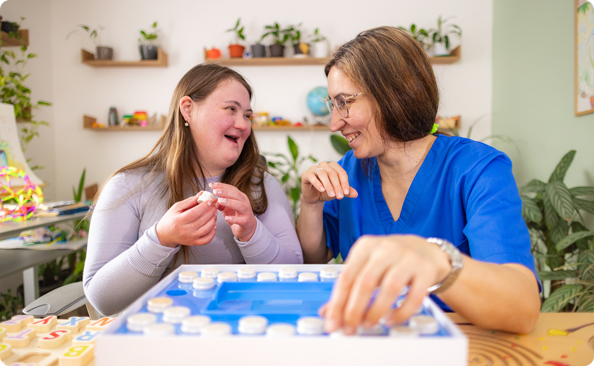 Two smiling women play a colorful board game together at a table in a bright, plant-filled room, sharing a warm, encouraging moment.
